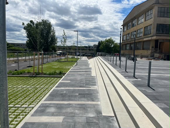 Rozsáhlá venkovní plocha s kombinací šedé a světlé dlažby, betonovými schodišťovými stupni a zatravňovacími dlaždicemi u budovy